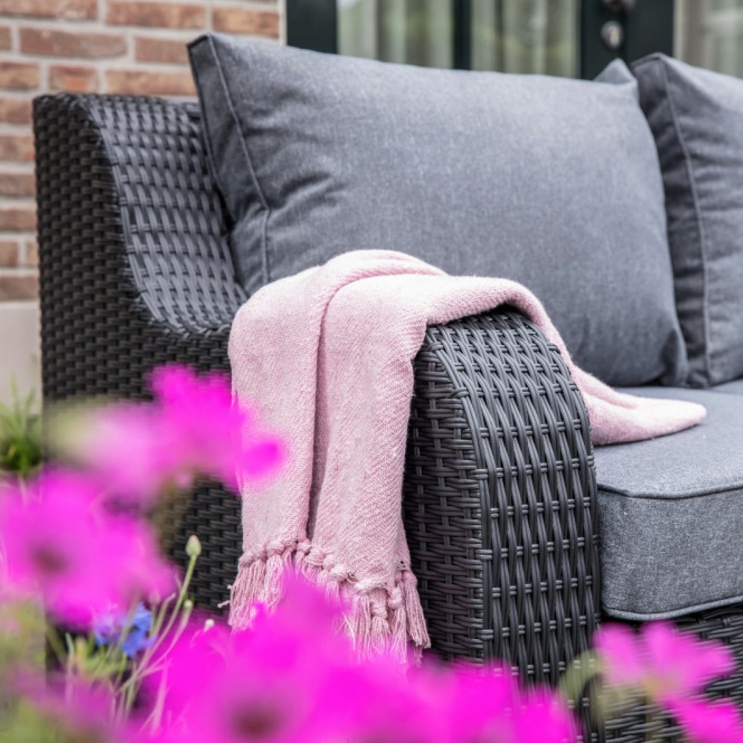 dunkelgraue Gartenmöbel mit bequemen Kissen und rosafarbener Decke; pinke Blumen im Vordergrund